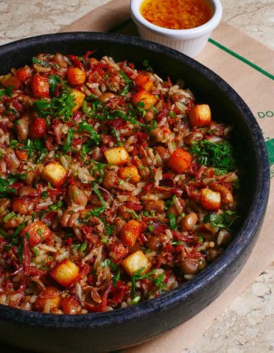 Baião de Dois served in a clay pot, made with carne de charque, rice, feijão de corda, bacon, and cubes of queijo coalho, accompanied by molho lambão on the side.