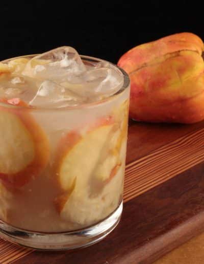 Glass of caipirinha de caju, filled with ice cubes and slices of cashew fruit, with a whole cashew fruit in the background, set on a wooden surface