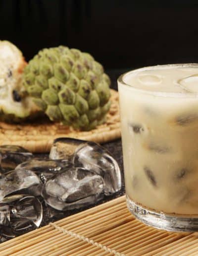 Caipirinha de fruta-do-conde, ao lado de uma fruta-do-conde aberta, com cubos de gelo espalhados sobre uma superfície texturizada.