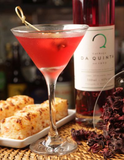 Pairing of dadinho with a red cocktail, alongside a glass of dried hibiscus flowers and a bottle of Cachaça Da Quinta in the background.