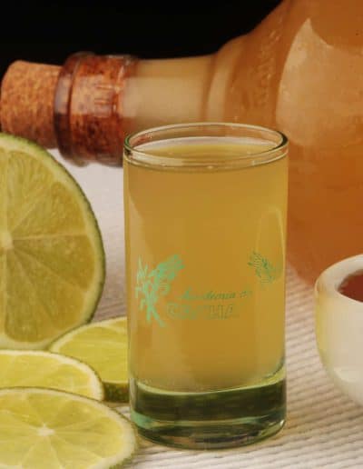 Image of an Academia da Cachaça glass with Melzinho, a golden drink made with aged cachaça and honey. Served with fresh lime slices, a bowl of honey, and a bottle of the product in the background, highlighting the fresh ingredients.